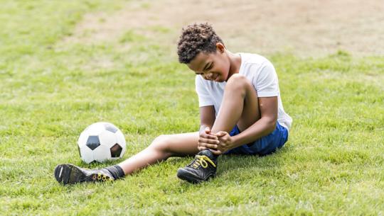 young boy holding ankle outside next to soccer ball